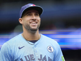 Portrait of a smiling baseball player wearing a light blue jersey and cap, standing in a stadium setting.