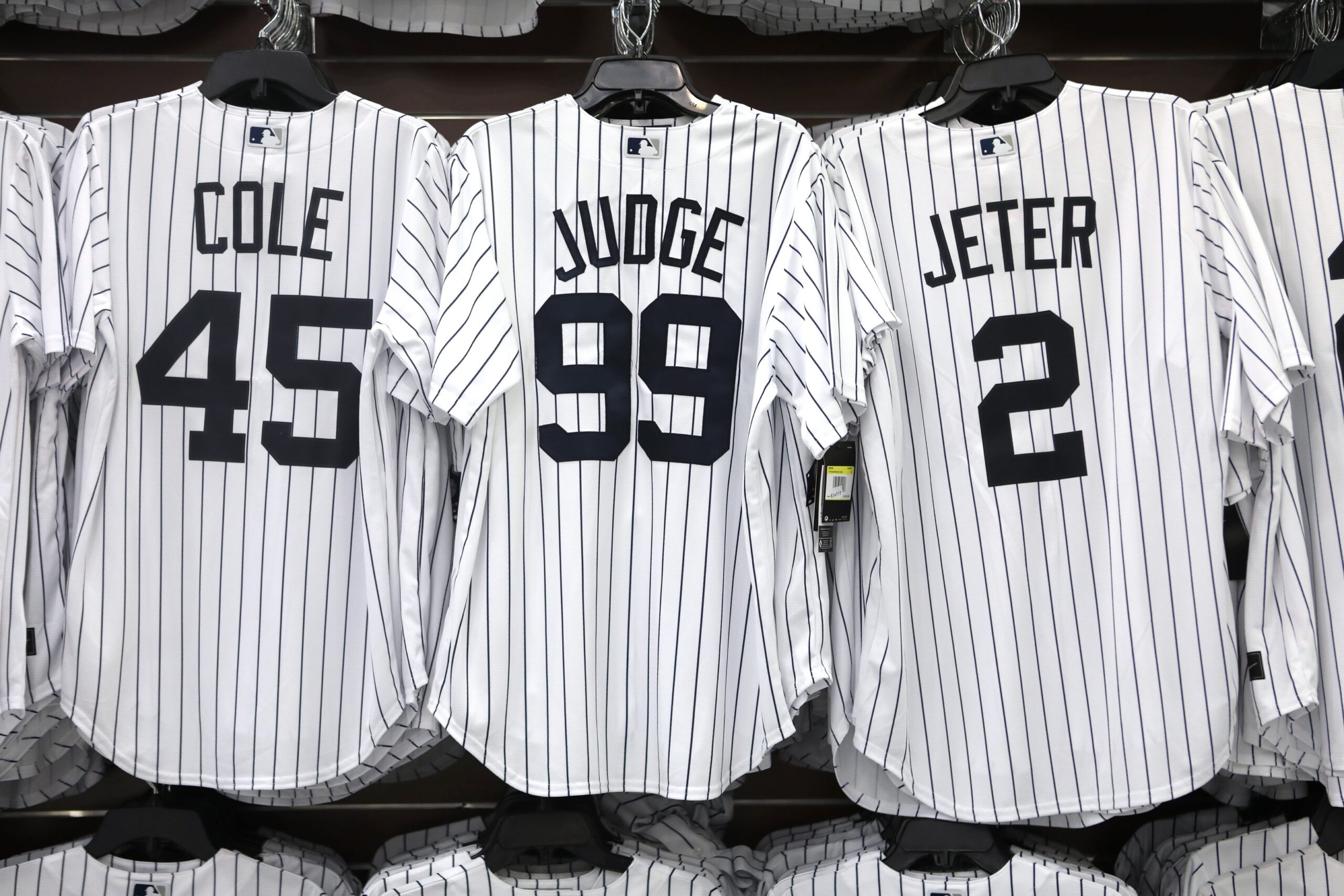 Row of white pinstripe baseball jerseys on a rack, showing names COLE (45) on the left and JETER (2) on the right, with a center jersey displaying number 99.