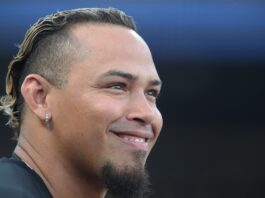 Close-up of a smiling man's face, ear with a silver hoop earring, short beard, and hair with blond tips against a dark background.