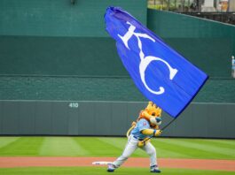 Mascot in blue uniform runs with a large blue flag bearing white lettering on a baseball field.
