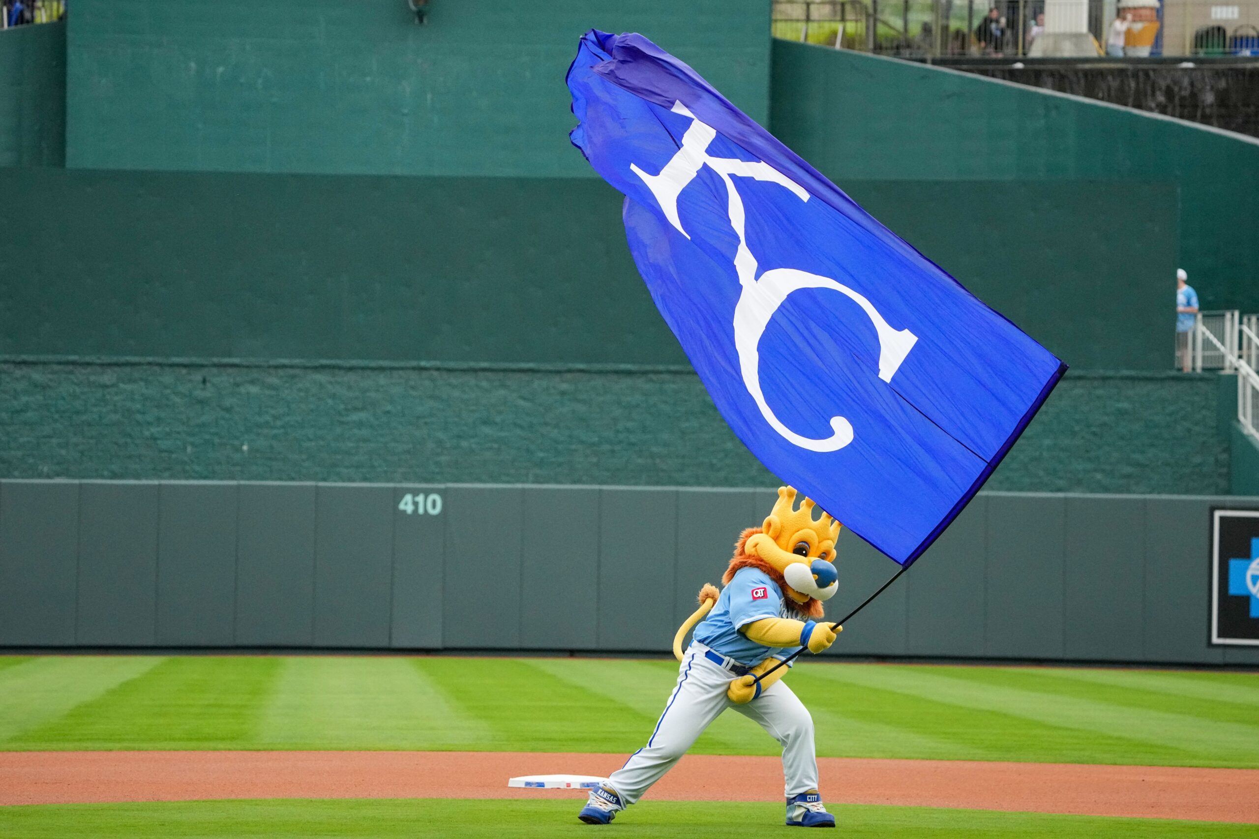 Mascot in blue uniform runs with a large blue flag bearing white lettering on a baseball field.