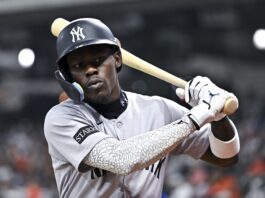 Baseball player in gray uniform swings a bat during a game, wearing a Yankees helmet and protective ear guard.