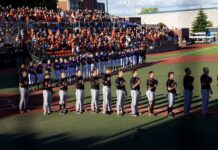 College Pitcher Suffers Serious Brain Injury In Car Crash Baseball players in purple jerseys and white pants stand in a line on the field as a large crowd watches from orange-seated stands behind them.