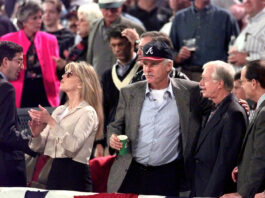 Crowd at a public event; a man in a baseball cap holds a green cup and chats with others nearby.