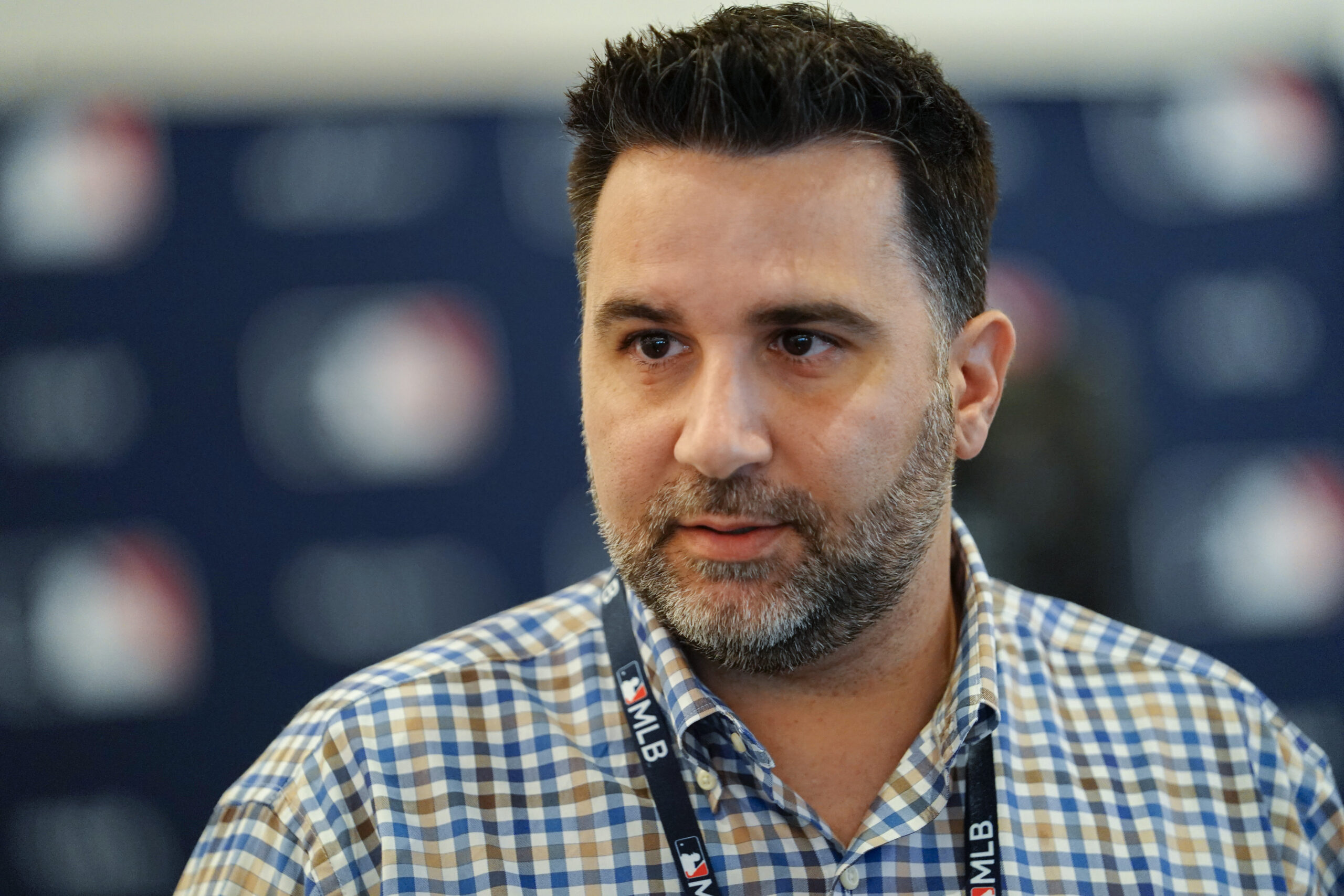 Close-up portrait of a man with dark hair and a gray beard, wearing a blue-checkered shirt and a lanyard, with a blurred background