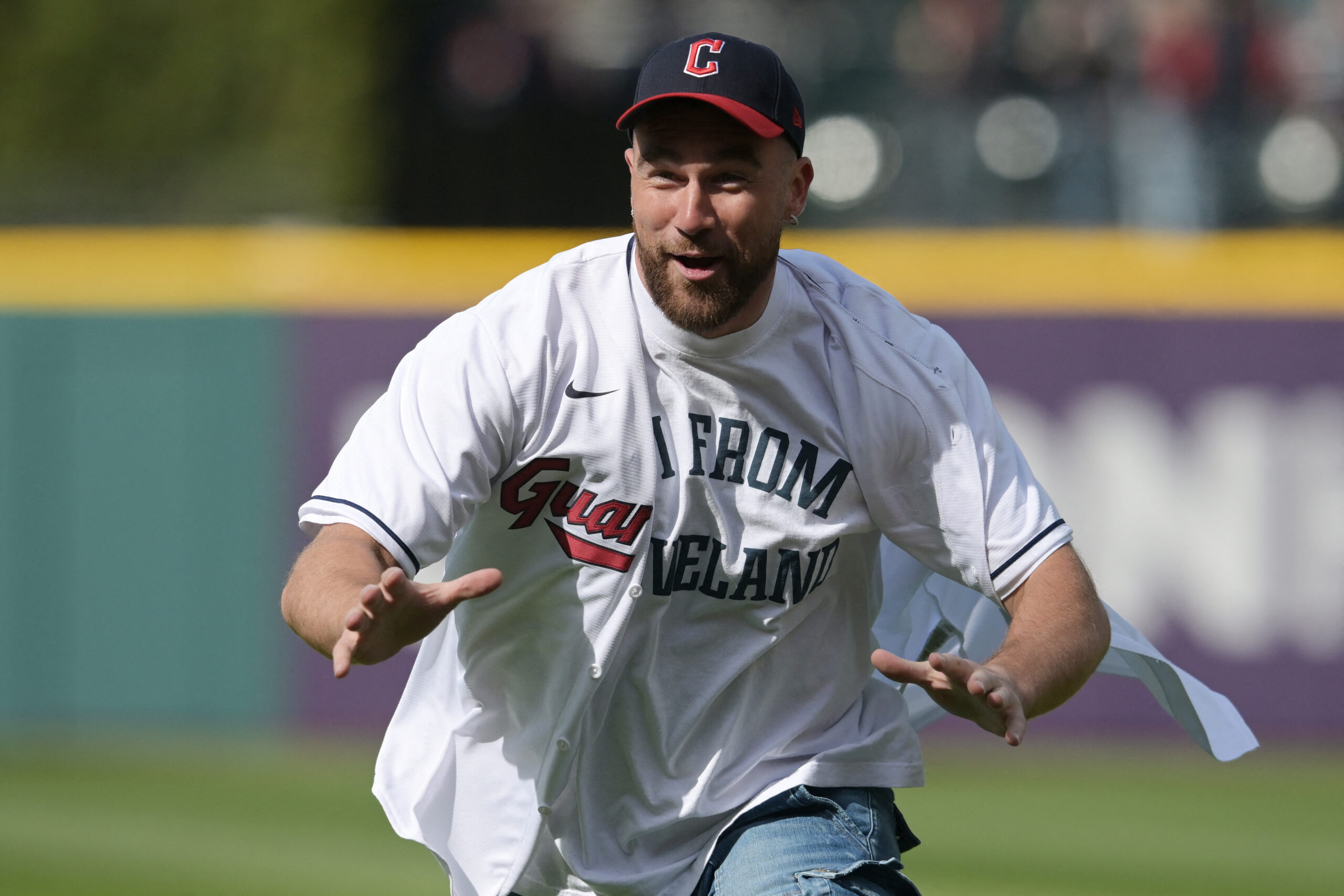 Travis Kelce Buys Massive Stake In His Beloved MLB Team Man in a white baseball jersey and jeans running toward the camera on a sunny field, wearing a dark cap with a red brim.
