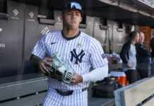 Yankees Star Is Rushed Off Field After Smashing His Face Baseball player in a Yankees pinstripe uniform holding a light-blue cleat in the dugout with teammates in the background