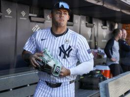 Baseball player in a Yankees pinstripe uniform holding a light-blue cleat in the dugout with teammates in the background