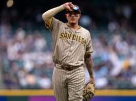 Baseball player in a tan pinstripe San Diego Padres uniform saluting with his right hand on a blurred crowd background, during a game.