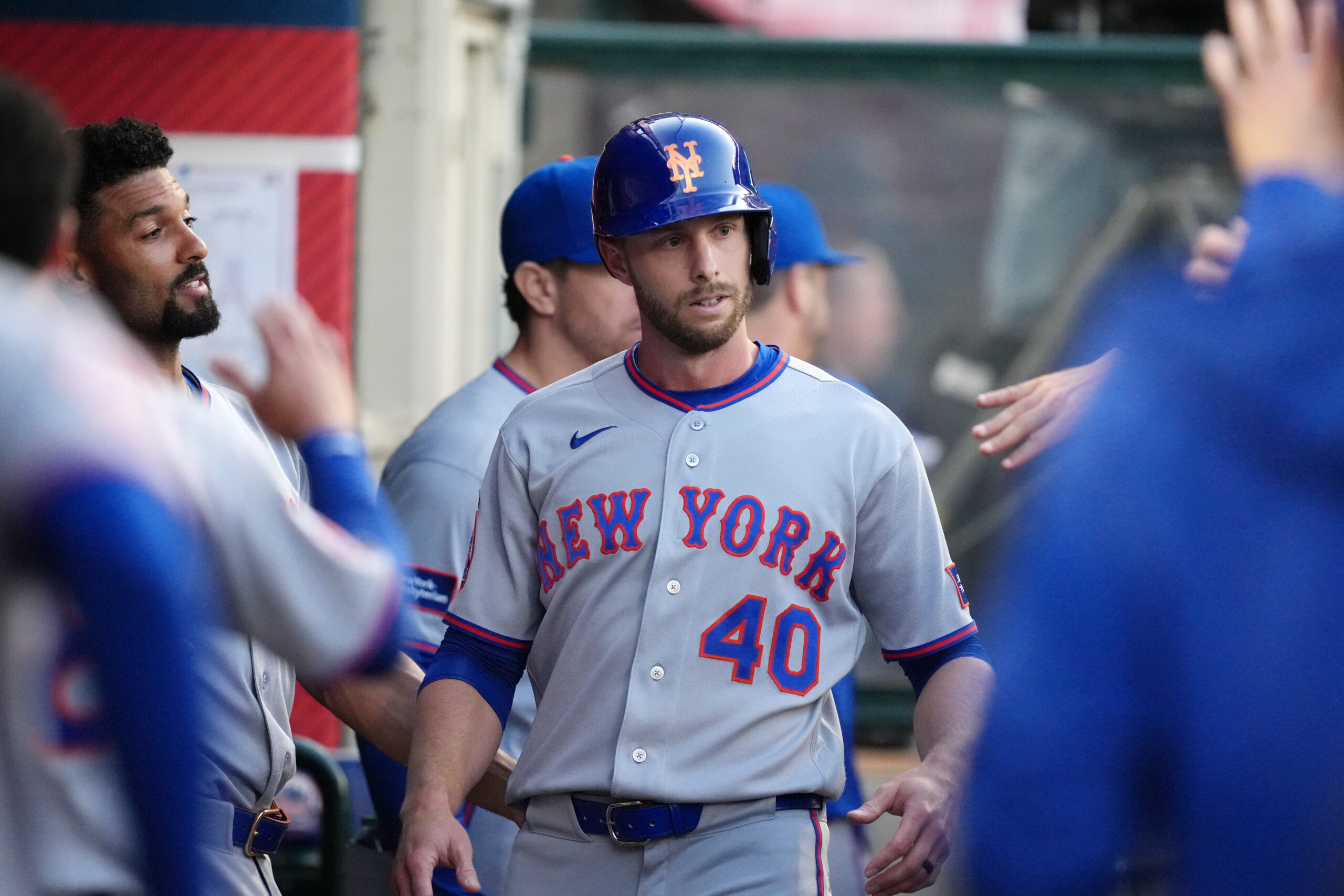 Baseball player in a gray New York Mets uniform wearing a blue helmet with an orange NY logo, standing in the dugout with teammates nearby.