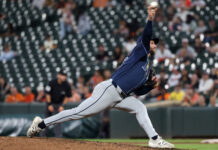 Gross Moment MLB Pitcher Throws Up On The Mound Baseball pitcher in a blue uniform delivering a pitch on the mound with blurred stadium crowd behind him.