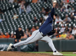 Baseball pitcher in a blue uniform delivering a pitch on the mound with blurred stadium crowd behind him.