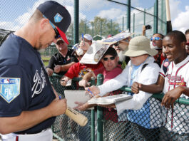 Baseball players sign autographs for fans pressed against a chain-link fence, pens and papers in hand.