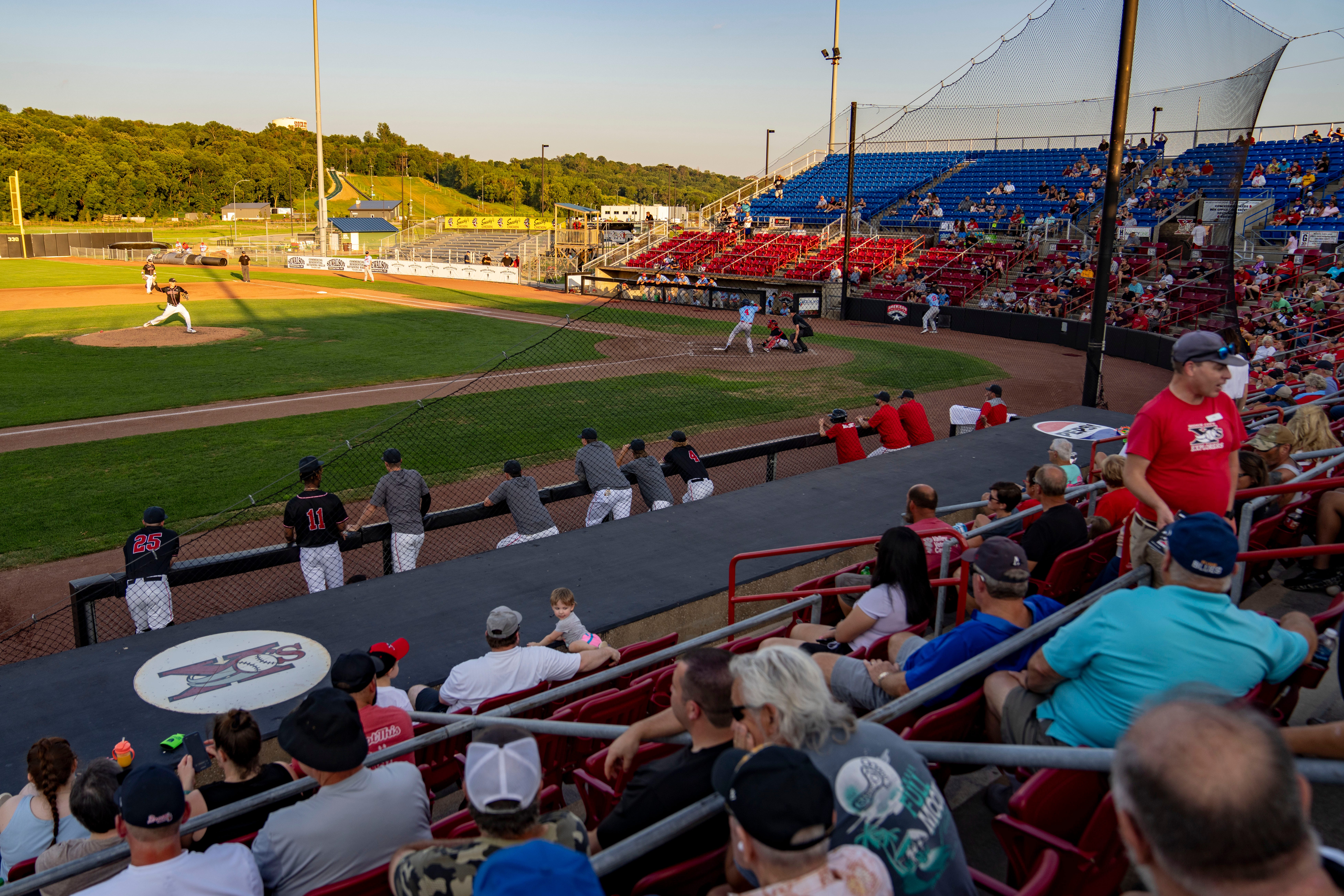 Baseball game in progress at a sunlit stadium, batter at plate and pitcher on mound, with audience in red and blue seats behind protective netting.