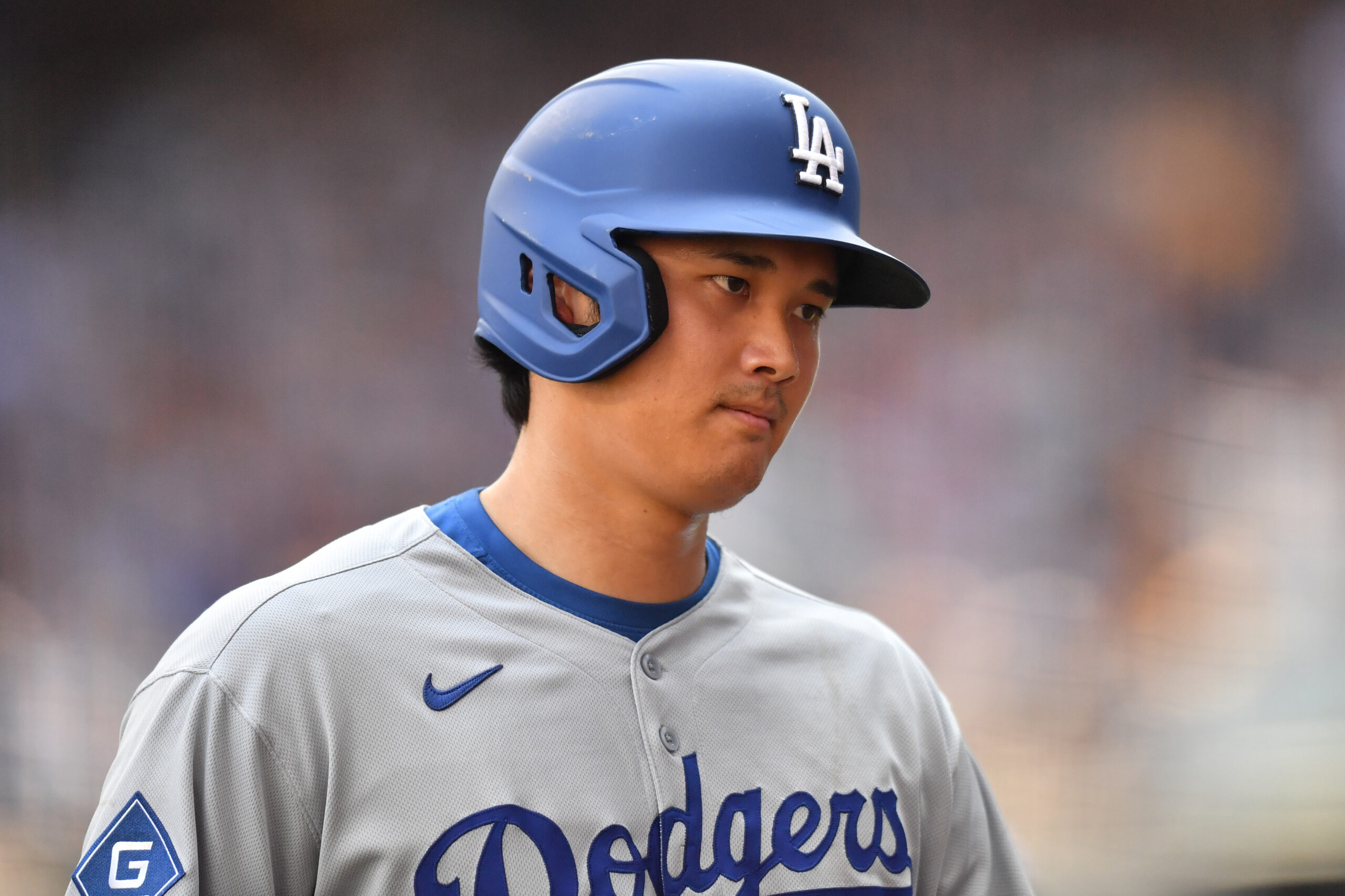 Baseball player wearing a blue Dodgers helmet and gray uniform, looking to the right with a blurred stadium background.