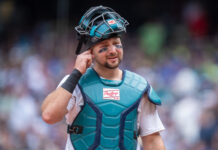 Mariners Announce Huge Cal Raleigh Injury Update Baseball catcher in teal protective gear touching his ear with one hand, crowd blurred in the background.