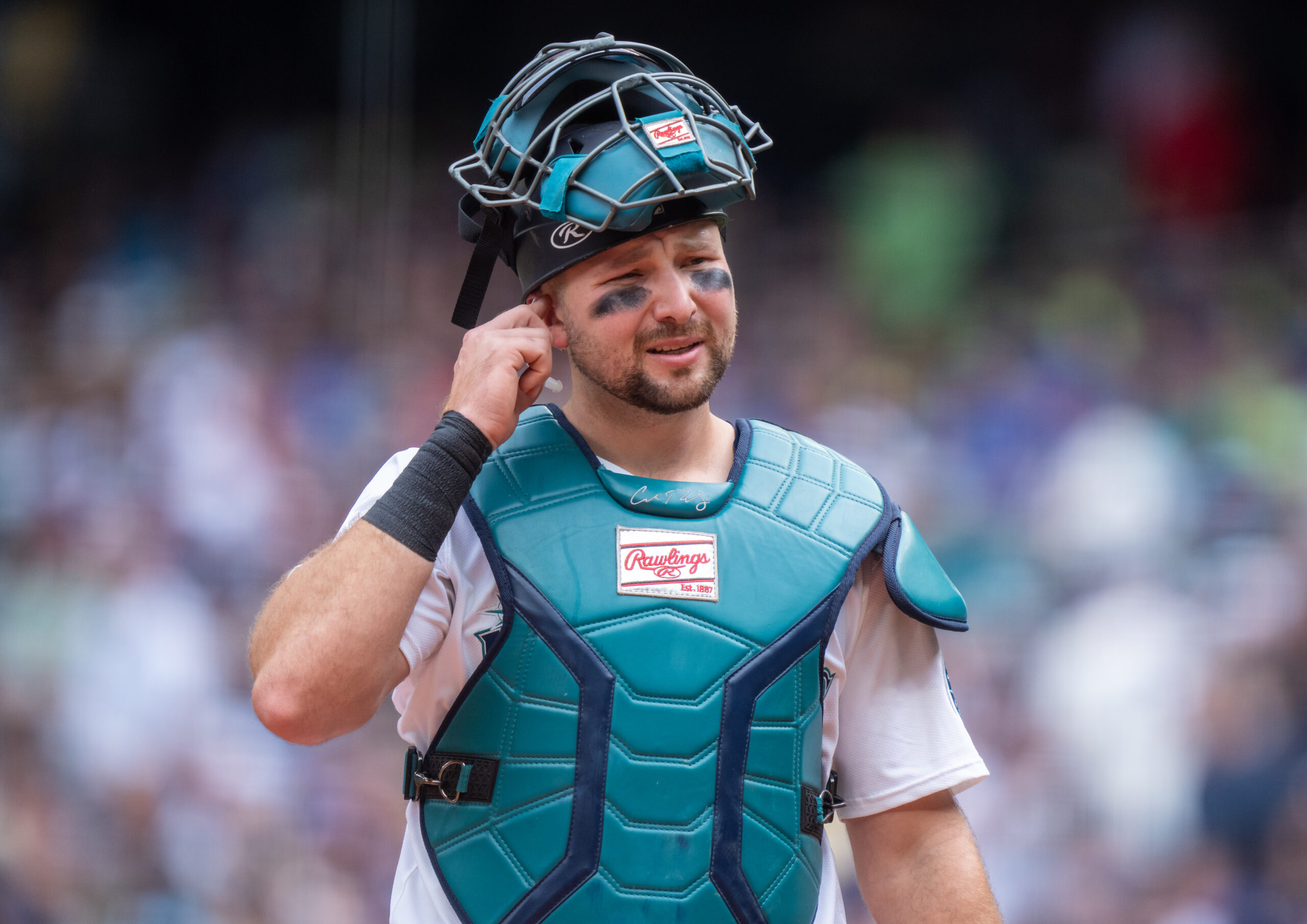 Mariners Announce Huge Cal Raleigh Injury Update Baseball catcher in teal protective gear touching his ear with one hand, crowd blurred in the background.