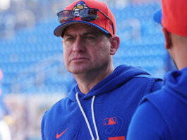Portrait of a man in a blue hoodie at a baseball stadium, orange cap with sunglasses perched on the brim, blue seats in the background kept out of focus.
