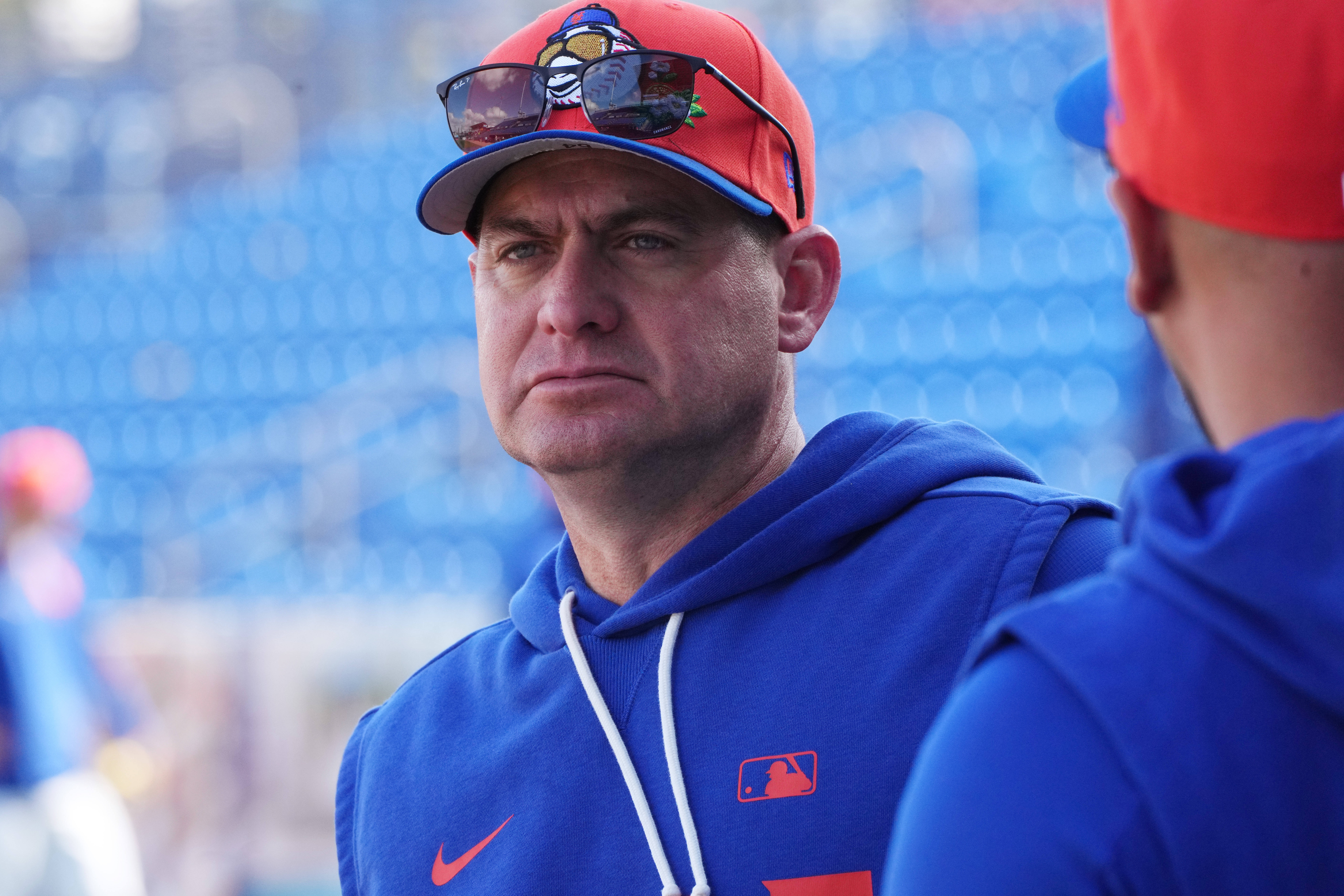 Portrait of a man in a blue hoodie at a baseball stadium, orange cap with sunglasses perched on the brim, blue seats in the background kept out of focus.