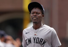 Yankees Livid After Star Man Plays With a Lollipop Baseball player in a gray New York jersey and black cap, smiling slightly outdoors.