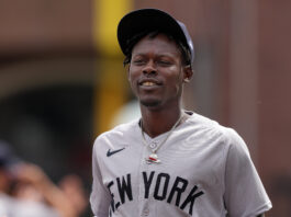 Baseball player in a gray New York jersey and black cap, smiling slightly outdoors.