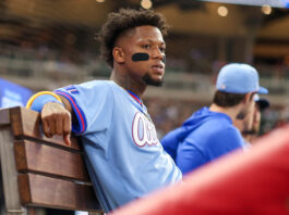 The Braves Pressure is Mounting as Injured List Grows Baseball player in a light-blue jersey sits on a wooden dugout bench with eye-black under his eyes, stadium lights blurred in the background.