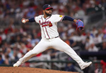 Braves Make Major Pitcher Move Amid Rotation Struggles Braves pitcher in white uniform mid-pitch on the mound, throwing toward home plate with a blue glove on the right hand and a crowd backdrop behind.