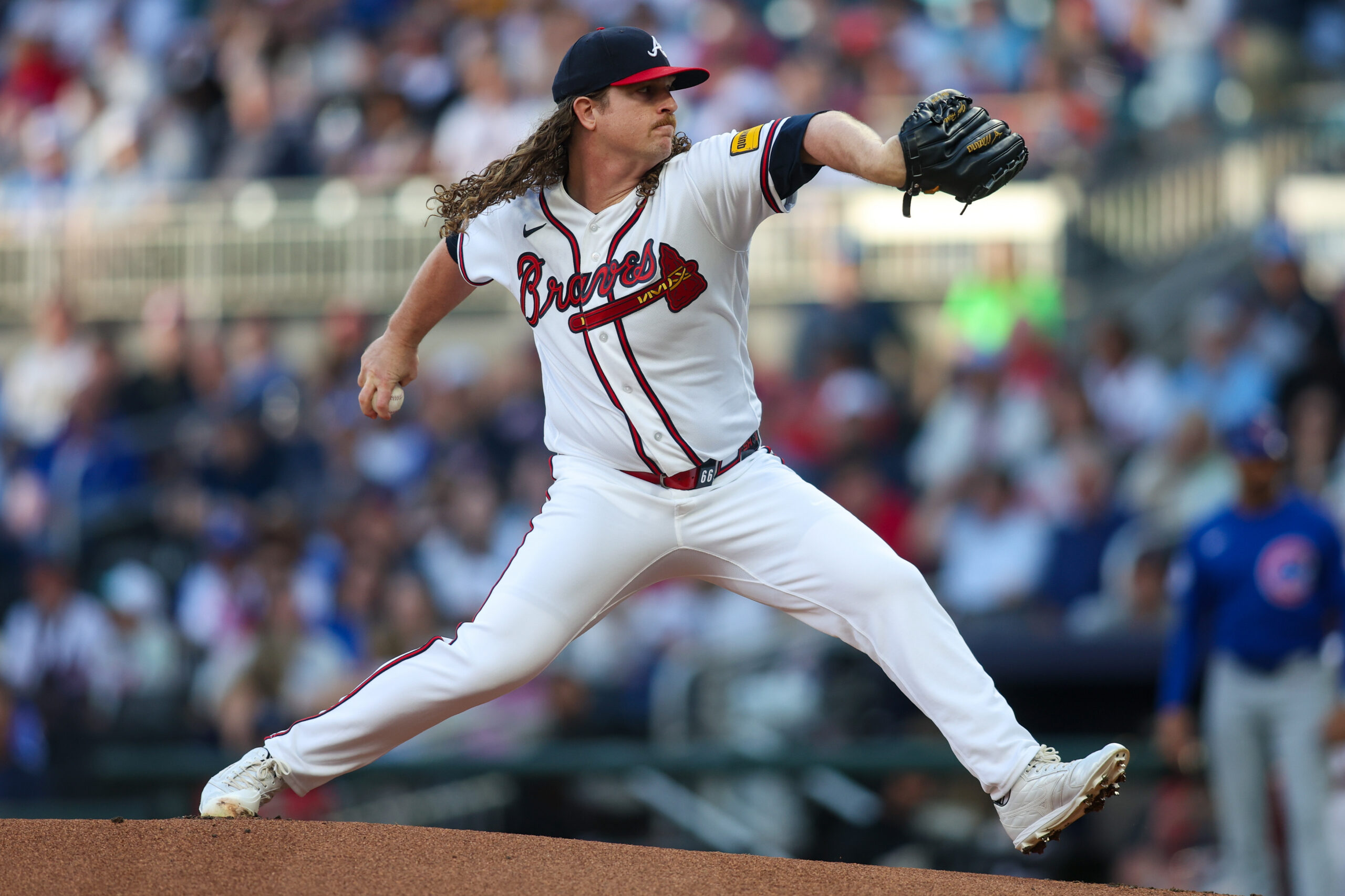 Baseball pitcher in a Braves uniform mid-pitch on the mound with a blurred crowd in the stands behind him.