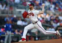Braves Lose Again As Lead Shrinks to 3 Points Baseball pitcher in a Braves uniform mid-pitch on the mound, red glove raised, with a blurred crowd in the background.