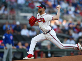 Baseball pitcher in a Braves uniform mid-pitch on the mound, red glove raised, with a blurred crowd in the background.
