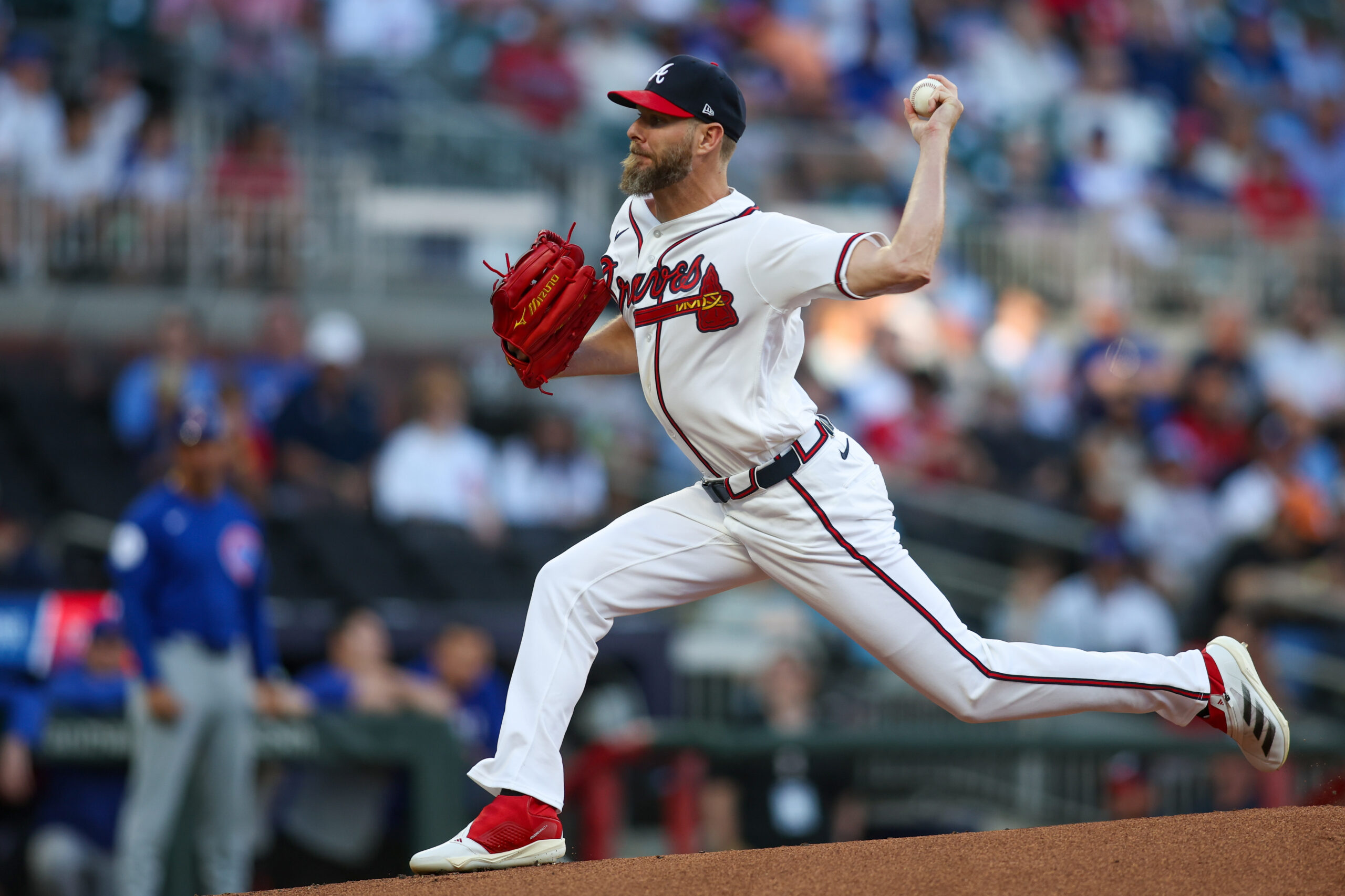 Baseball pitcher in a Braves uniform mid-pitch on the mound, red glove raised, with a blurred crowd in the background.