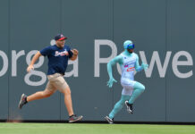 Atlanta Braves Fan Taunts Popular Mascot Two men sprinting side by side along a ballpark outfield wall; one wears a navy Braves shirt, beige shorts, and a cap, the other in a turquoise bodysuit with goggles.