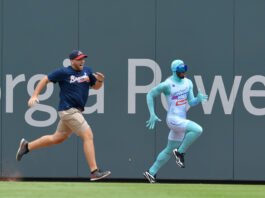 Two men sprinting side by side along a ballpark outfield wall; one wears a navy Braves shirt, beige shorts, and a cap, the other in a turquoise bodysuit with goggles.