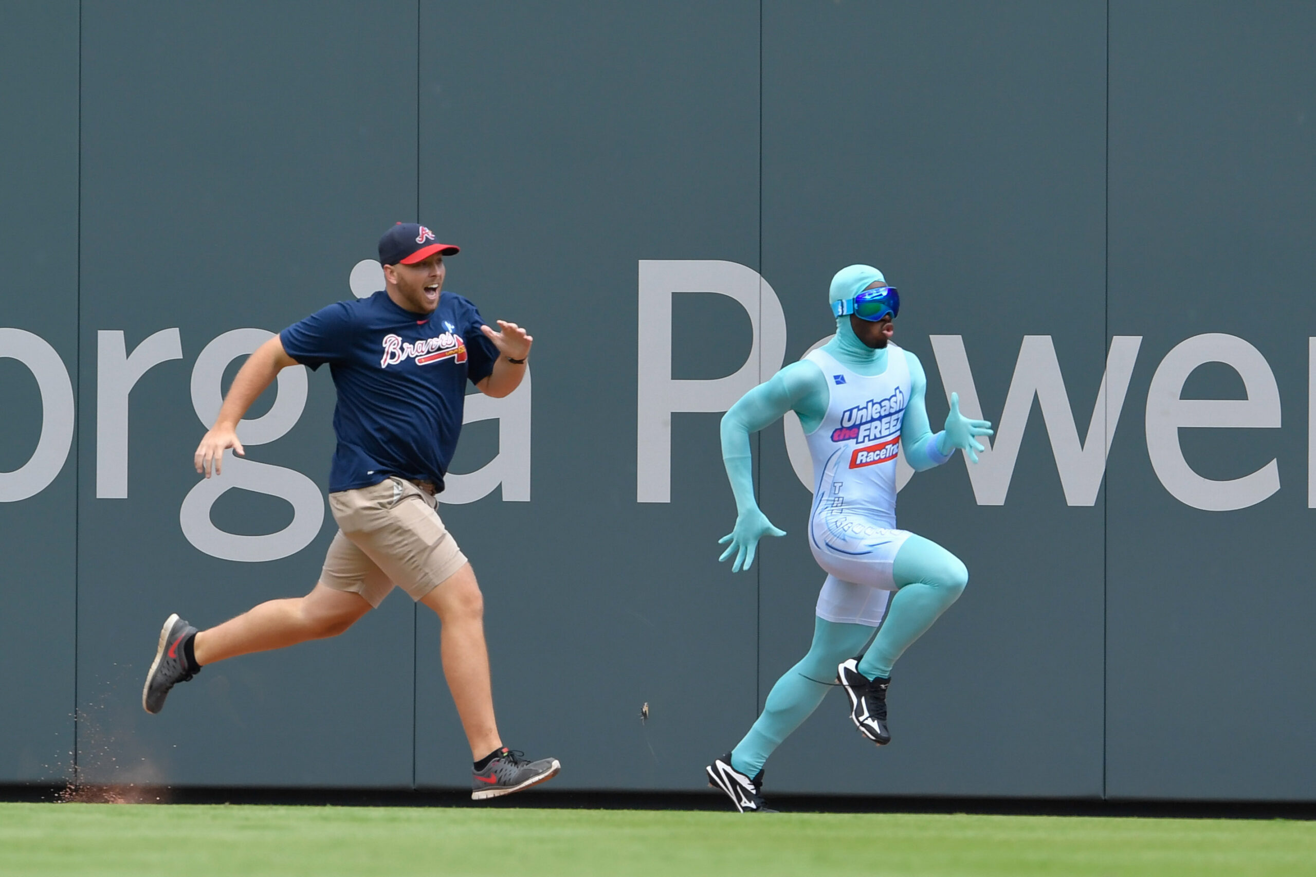 Two men sprinting side by side along a ballpark outfield wall; one wears a navy Braves shirt, beige shorts, and a cap, the other in a turquoise bodysuit with goggles.