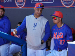 Baseball players in Mets uniforms stand in the dugout, one gesturing while another wears orange cap and reflective sunglasses, behind a blue padded railing.
