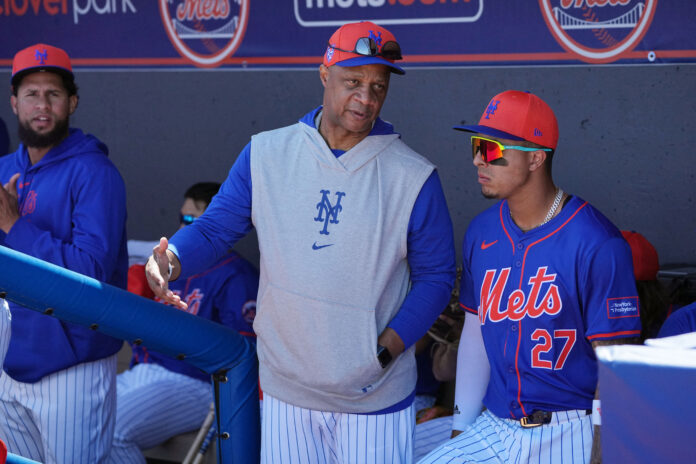 Baseball players in Mets uniforms stand in the dugout, one gesturing while another wears orange cap and reflective sunglasses, behind a blue padded railing.