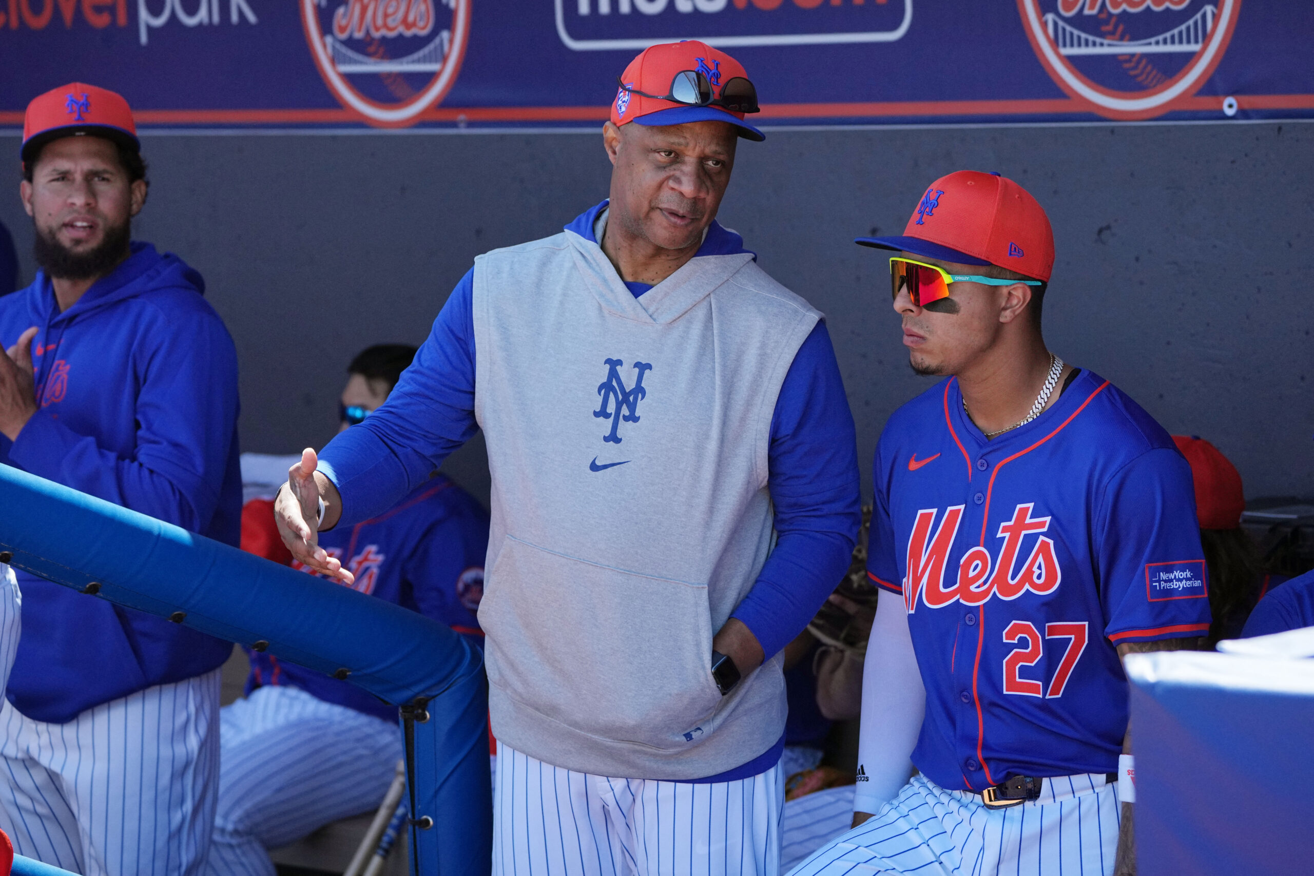 Baseball players in Mets uniforms stand in the dugout, one gesturing while another wears orange cap and reflective sunglasses, behind a blue padded railing.