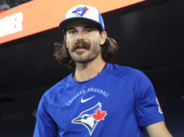 Man with long hair and a mustache wearing a blue Toronto baseball shirt and a Toronto Blue Jays cap, standing in a dark setting with a red banner behind him.