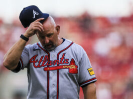 Baseball coach in Braves uniform tipping his cap while looking down on a field with red stadium seats in the blurred background