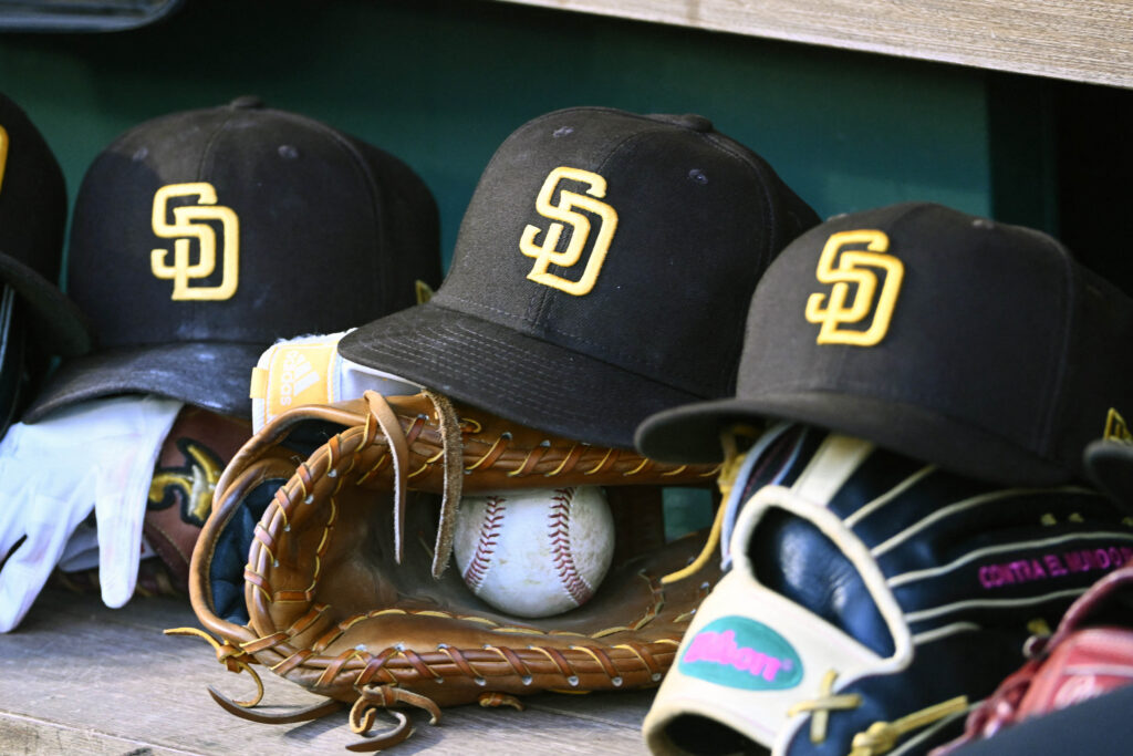 Stack of black baseball caps with yellow SD logos, a baseball, and a brown glove on a shelf, cluttered with other gear.