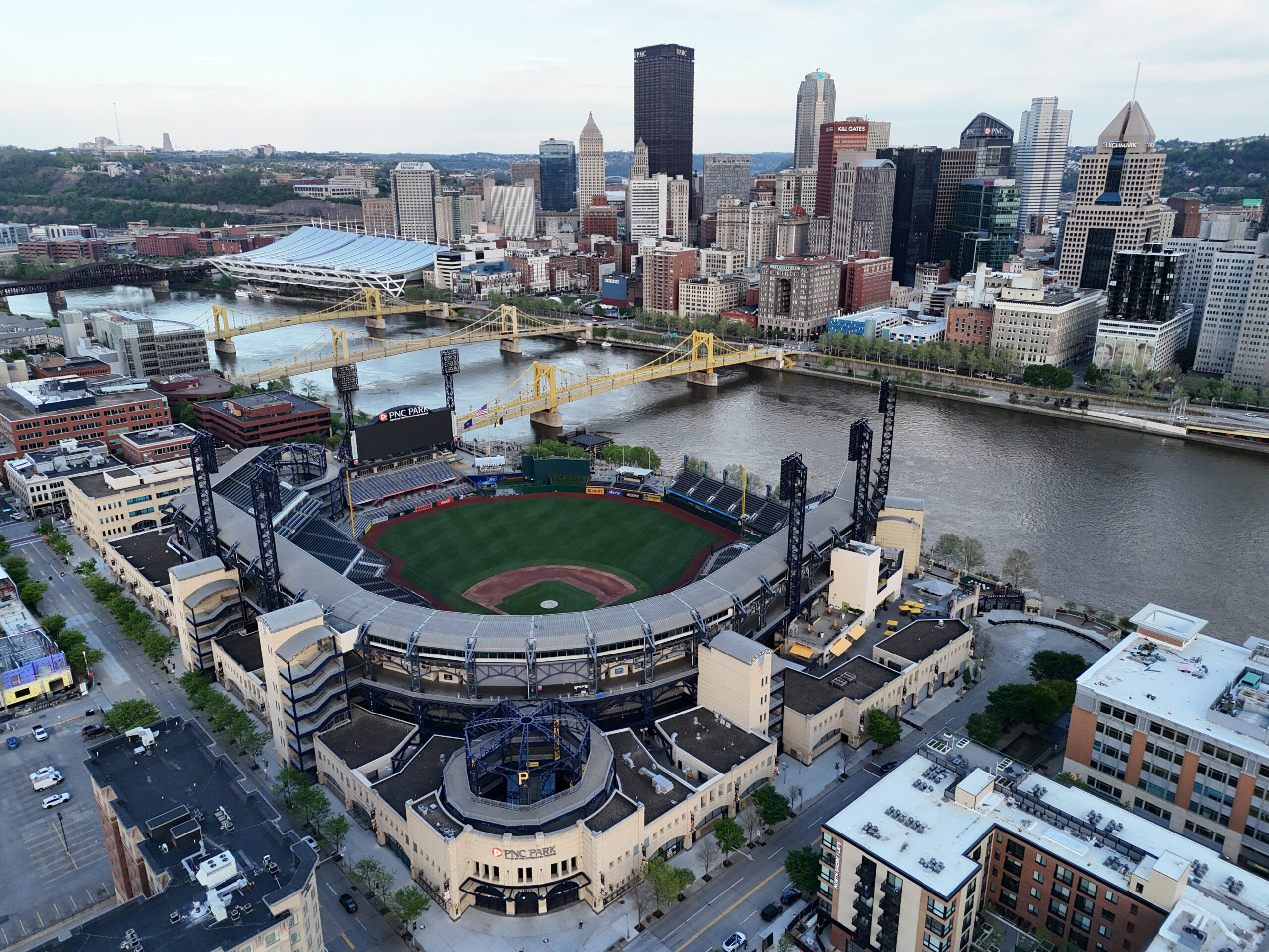 Aerial view of Pittsburgh with the U.S. Steel skyline, the Allegheny River, and PNC Park in the foreground beneath a cloudy sky.