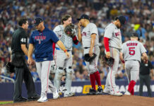 Little-Known Rule Sparks Chaos in Red Sox-Blue Jays Clash Group of Boston Red Sox players and an umpire standing on a pitcher's mound during a game, with a crowded stands in the background