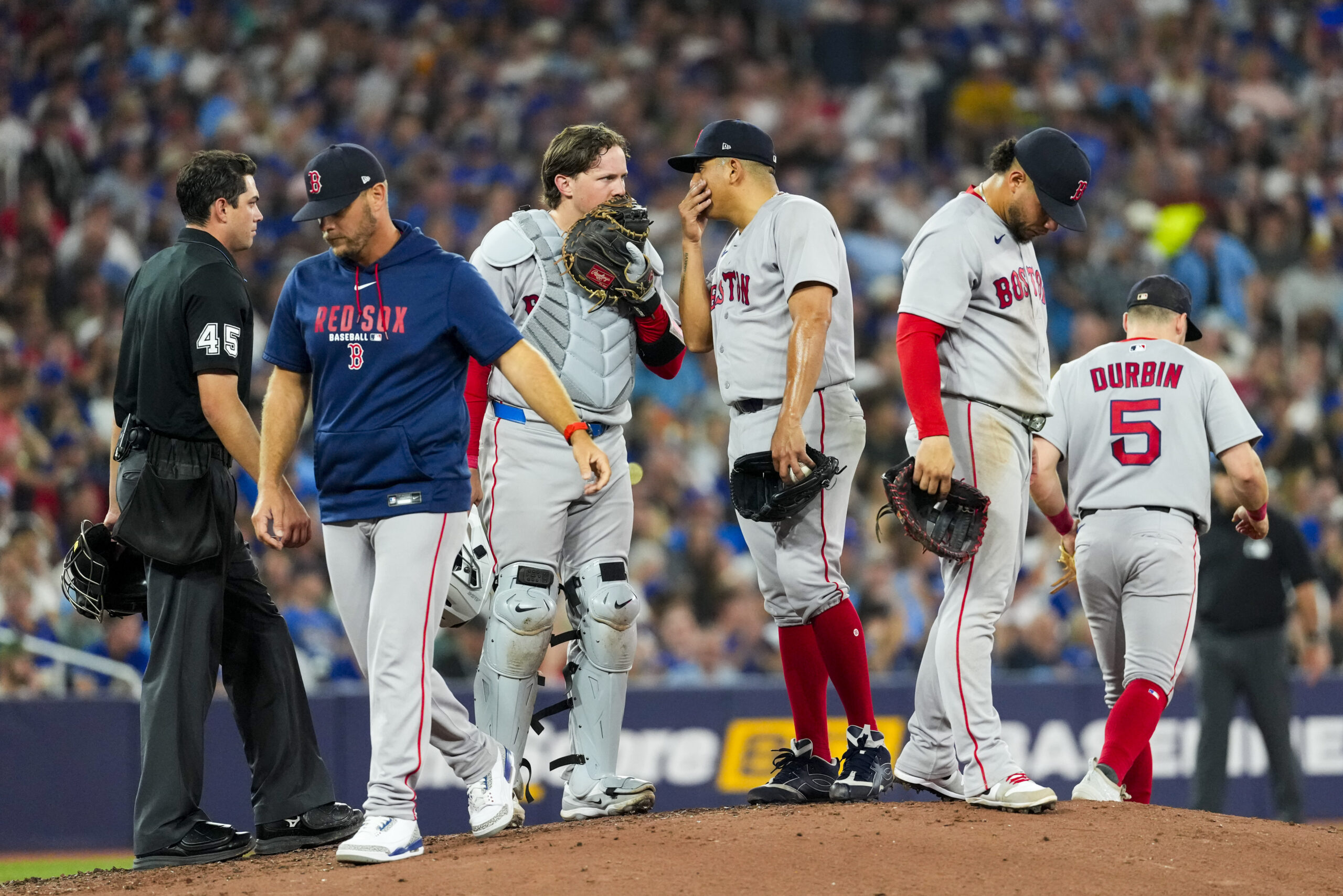 Group of Boston Red Sox players and an umpire standing on a pitcher's mound during a game, with a crowded stands in the background